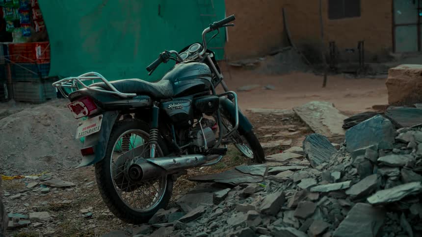 A weathered black motorcycle stands parked on uneven, rocky ground in a rustic, rural location