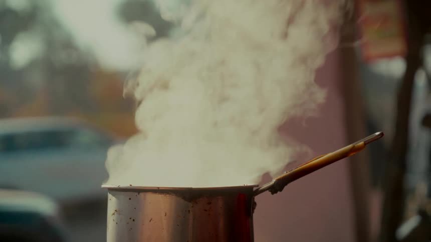 A close-up slow-motion shot of a person preparing traditional Indian chai tea in a steaming pot at a roadside stall