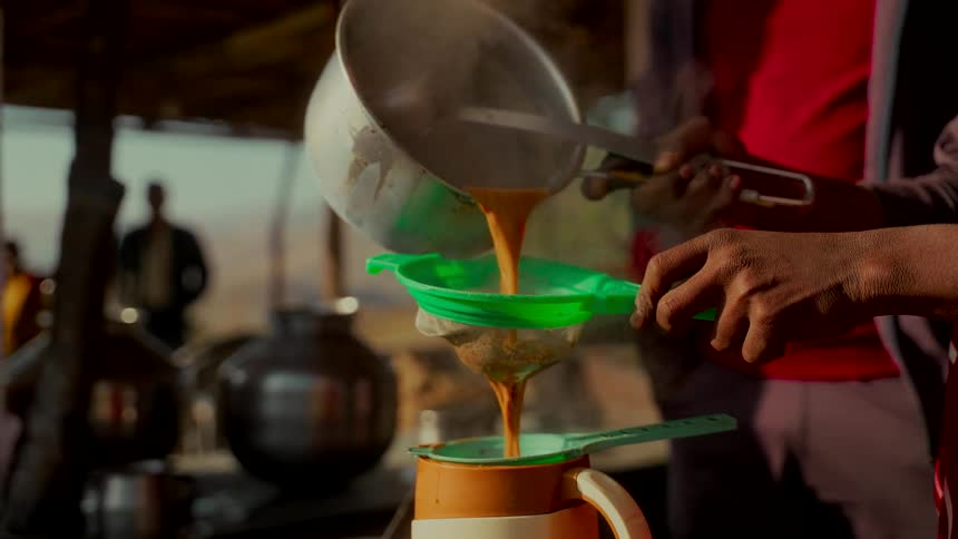 A close-up slow-motion shot shows a person straining hot, steamy Masala Chai tea into a jug