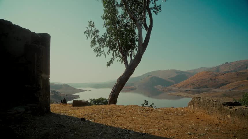 A peaceful landscape view of a tranquil lake nestled in dry, golden hills, framed by a lone tree and ancient ruins