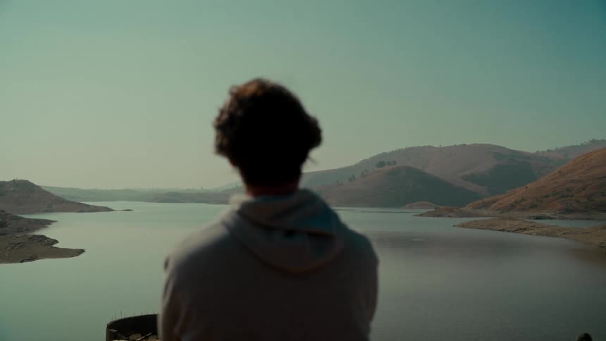 Static shot from behind a person in a hoodie, looking out over a calm lake nestled between dry, rolling hills