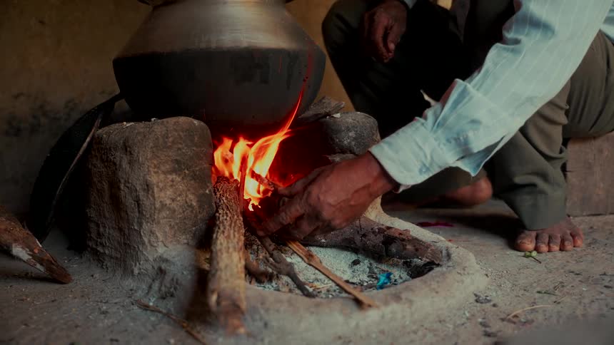 A man's hands stoke a traditional clay cooking fire beneath a large simmering pot in a rustic setting