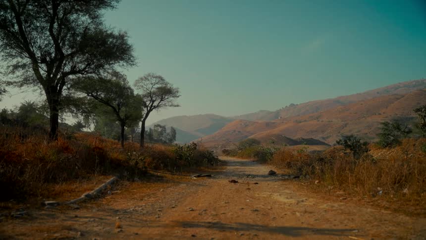 A quiet, dusty road winds through an arid landscape of rolling brown hills under a clear sky