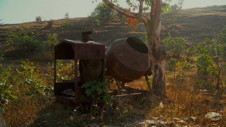 An old, rusted concrete mixer sits abandoned and overgrown in a sunlit, rural field