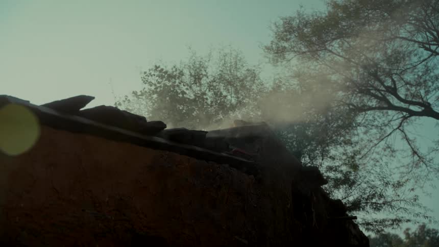 Smoke billows from the rooftop of a rustic mud house as the sun shines through the trees