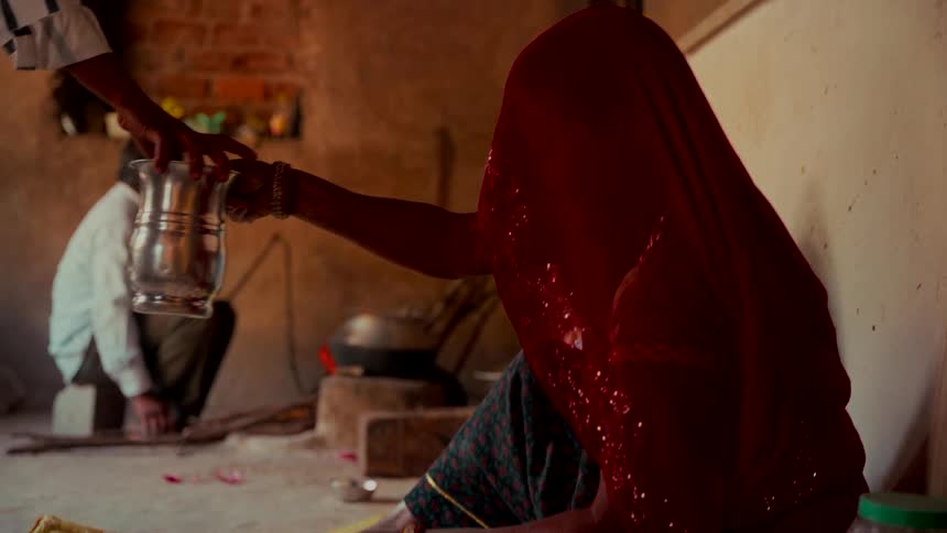 A woman in a traditional red Indian sari kneads dough on the floor of a rustic home as someone hands her a metal water vessel