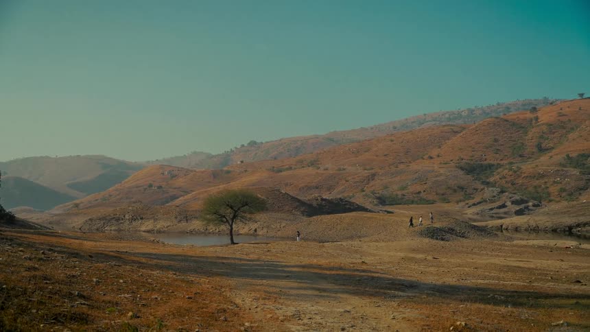A serene, wide shot shows a vast, arid landscape with rolling brown hills, a solitary tree by a small pond, and distant figures walking under a clear sky