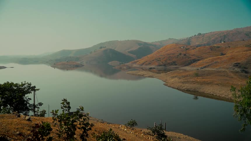 A serene and expansive view of a calm lake reflecting the arid, rolling hills under a hazy, clear sky