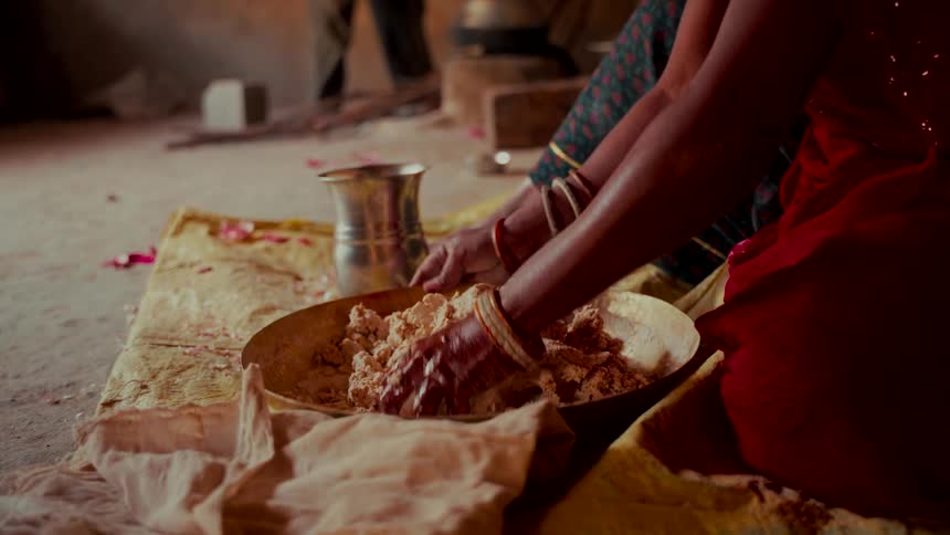 An Indian woman in a red sari kneads dough in a brass bowl while sitting on the floor in slow motion
