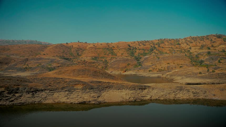 A static shot captures the serene beauty of a calm lake and arid, orange hills under a vast, clear teal sky