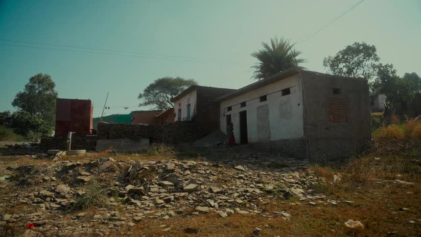A rustic village home stands amidst a rocky, sun-drenched landscape under a clear sky