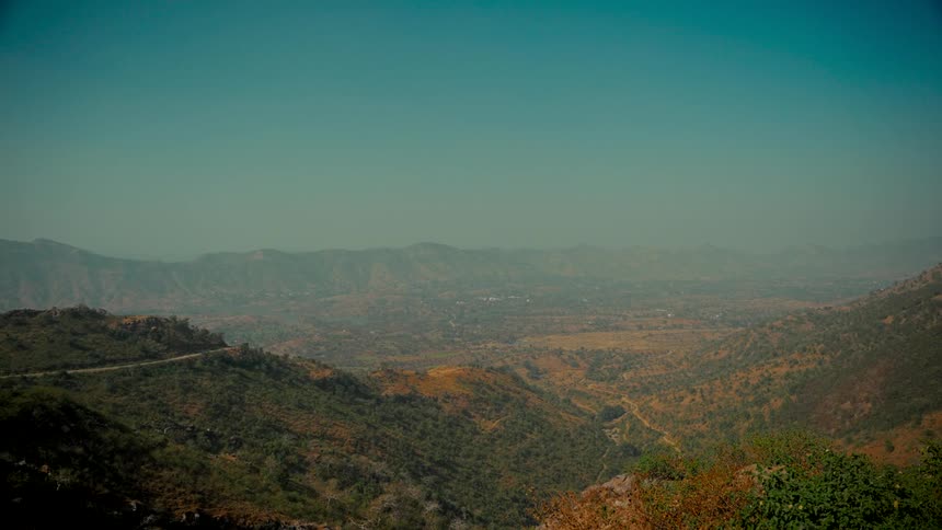 A wide, static shot captures a vast and hazy mountain valley on a clear day, showcasing the arid landscape and a winding road