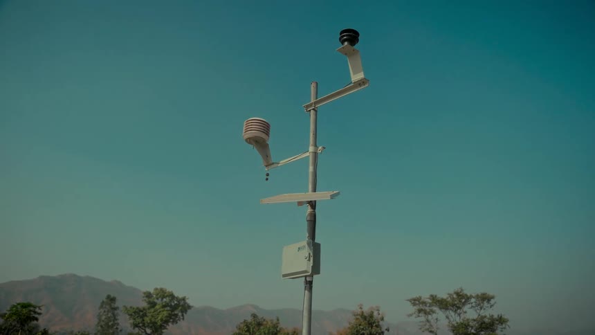 A remote weather station with various sensors monitors atmospheric conditions against a clear blue sky with mountains in the background