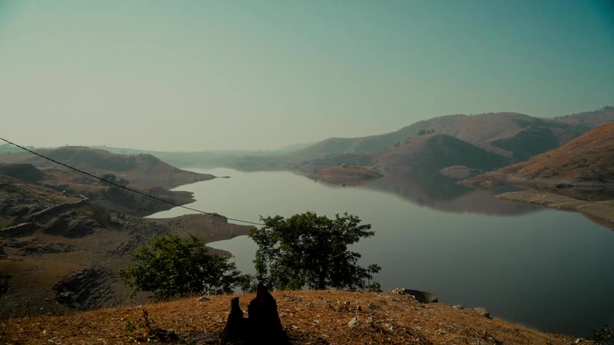 A tranquil lake sits amidst a vast, arid landscape of rolling hills, with the camera slowly pulling back to reveal the expansive scenery