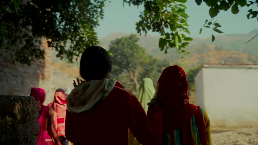 A group of Indian women in colorful traditional clothing walk together through a rural village in slow motion