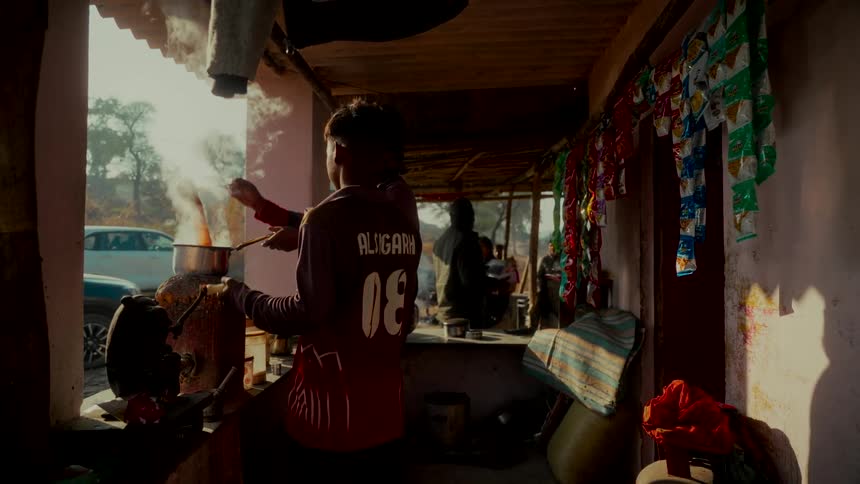 A young man wearing a jersey prepares a steaming meal at a rustic roadside food stall in India