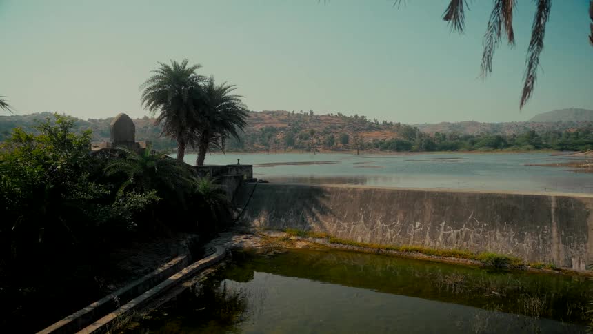 A tranquil wide shot of a concrete dam on a calm lake in a rural, sun-drenched landscape with palm trees