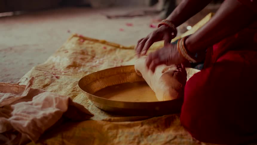 A woman wearing a red sari and bangles kneads dough in a traditional brass bowl on the floor