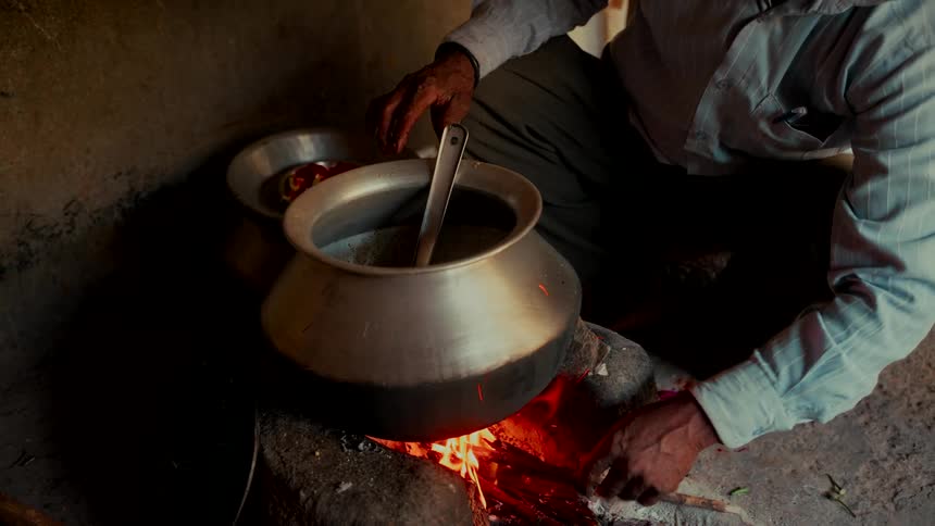 A man tends to a fire while cooking a meal in a large pot over a traditional chulha stove