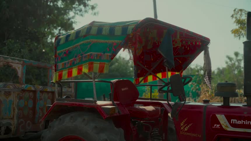 A red tractor with a colorful, traditionally decorated canopy and painted trailer is parked in a rural village