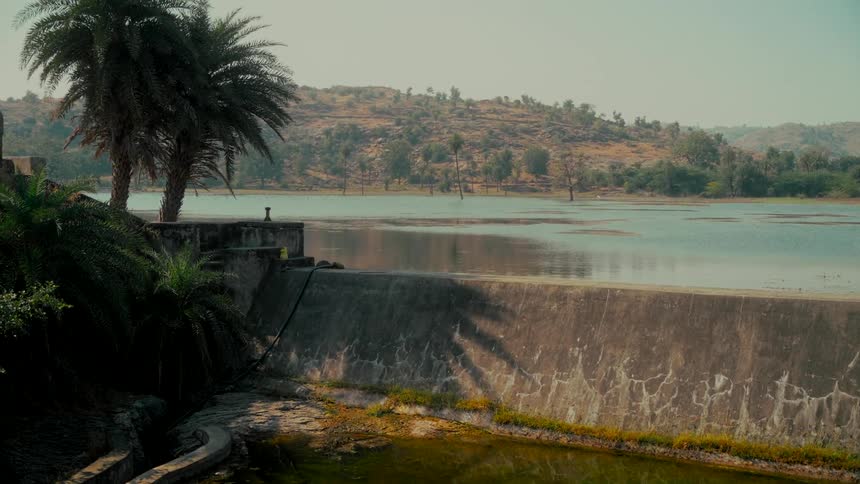 A serene view of a concrete dam holding back a tranquil lake surrounded by arid hills and palm trees