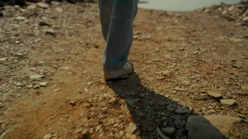 A person's legs and feet in jeans and sneakers are filmed from behind while walking in slow motion on a dusty, rocky path on a sunny day