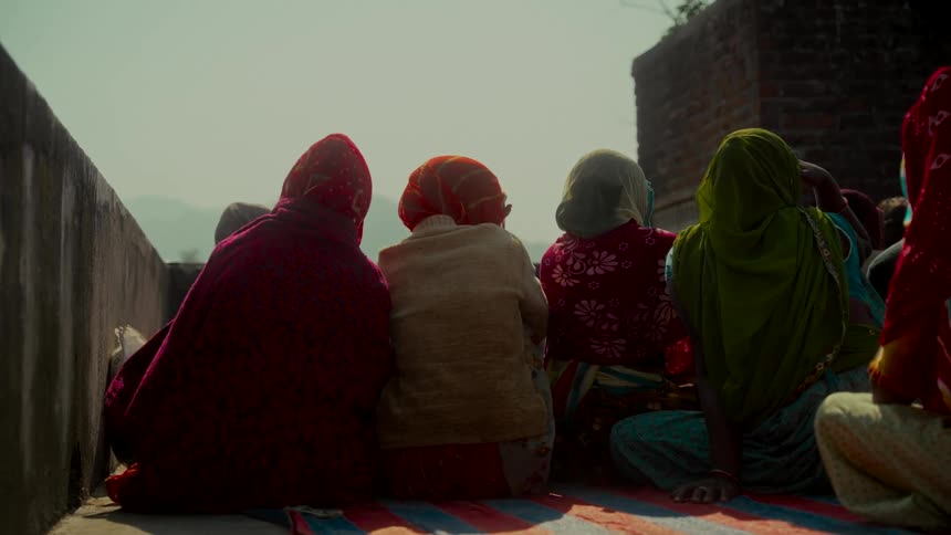 Several Indian women wearing colorful headscarves sit together on a striped mat on a sunny rooftop