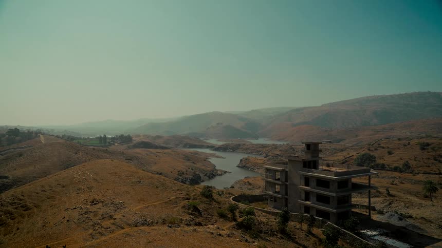 A wide static shot shows an unfinished building overlooking a winding river in a vast, arid, and hilly landscape