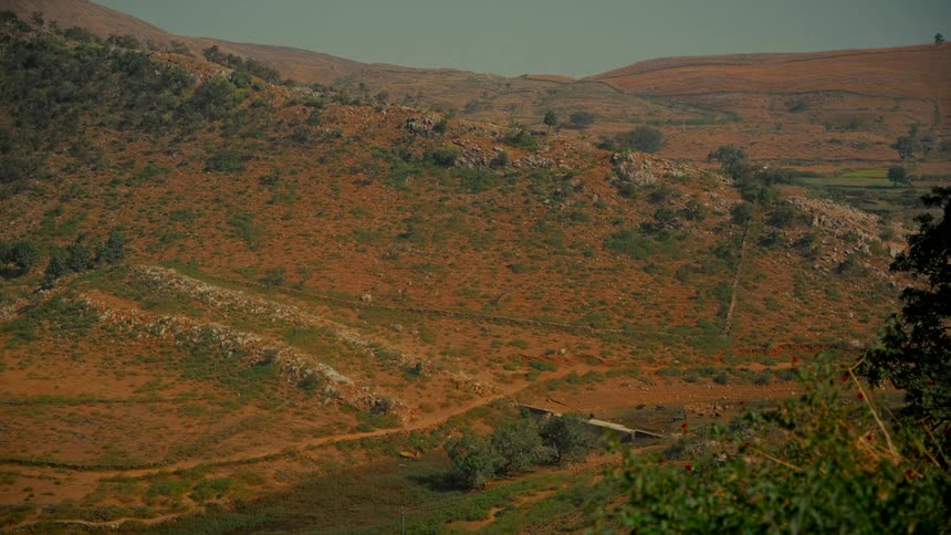 A static wide view of an arid, hilly landscape with orange soil, sparse vegetation, and winding dirt paths under a hazy sky