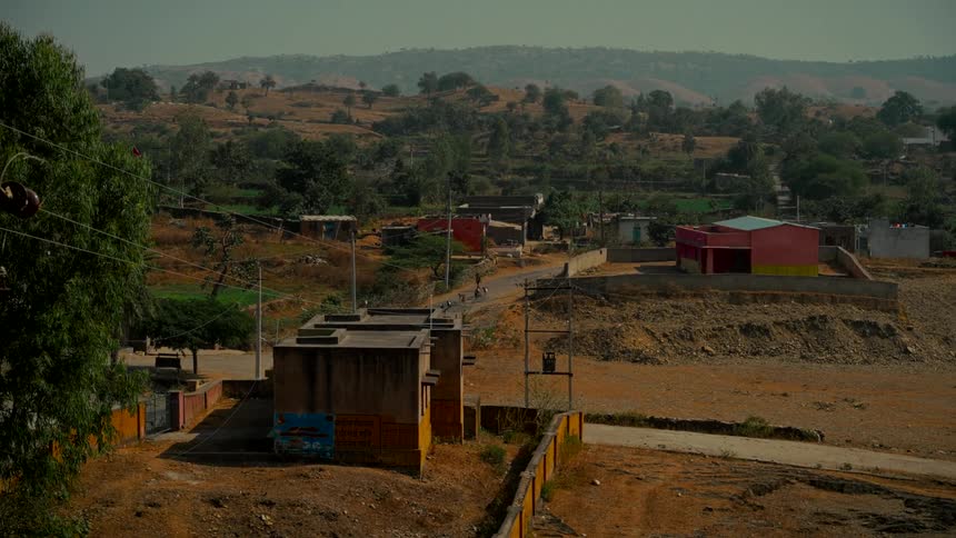 A wide, static shot of a quiet rural village in a dry, hilly landscape as a motorcycle drives down a distant road