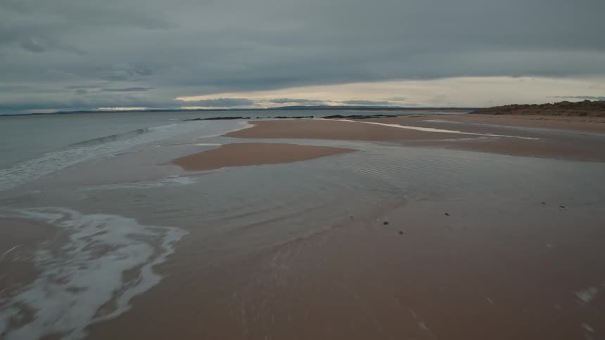 A serene aerial view flies low along the shoreline of a vast beach as gentle waves wash ashore on an overcast day
