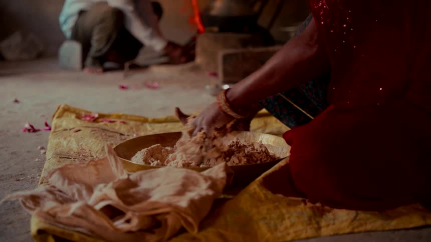 An Indian woman in a traditional red sari kneels on the floor, kneading dough in a brass bowl inside a rustic home