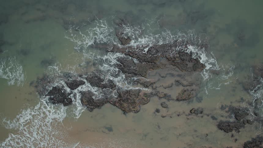 A top-down aerial shot shows ocean waves slowly breaking over large rocks on the shore