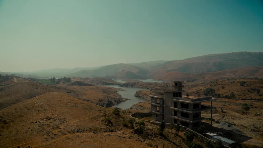 A desolate, unfinished building stands alone in a vast, arid landscape next to a winding river