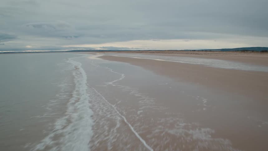 A low aerial shot flies along a vast, empty sandy beach as calm waves lap the shore under a moody, overcast sky