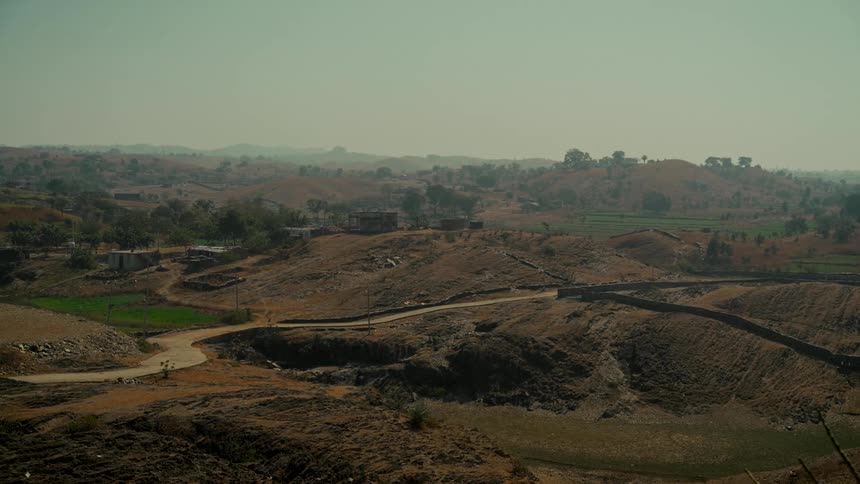 A static wide shot of a dry, hilly rural landscape with a winding road under a hazy sky