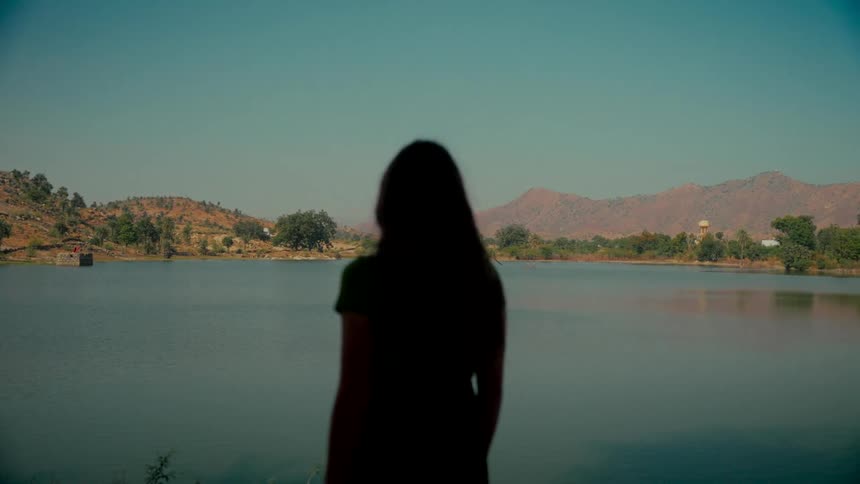 A silhouetted woman stands peacefully, looking out at a serene lake surrounded by rolling hills under a clear blue sky