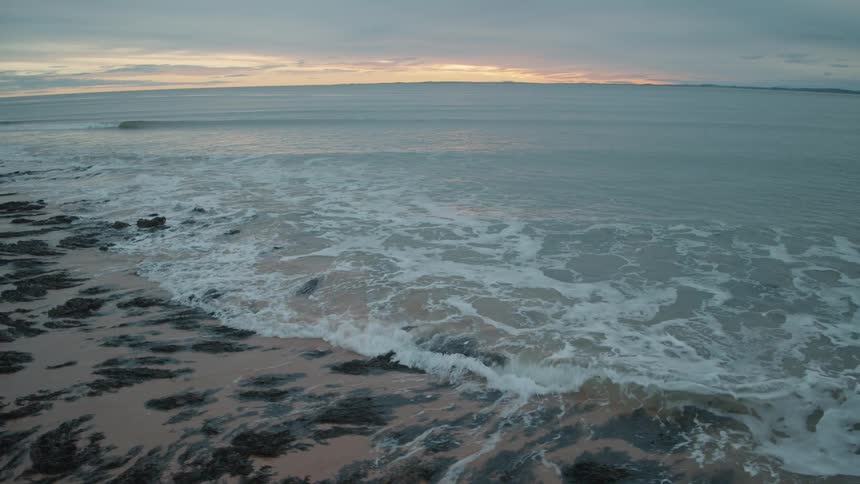 A gentle ocean wave rolls in and washes over the rocky, seaweed-covered shore in slow motion during a beautiful, cloudy sunset