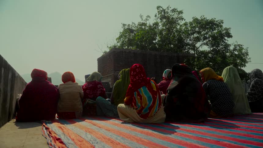 A group of Indian women in colorful traditional sarees sit together on a striped rug on a rooftop during a sunny day