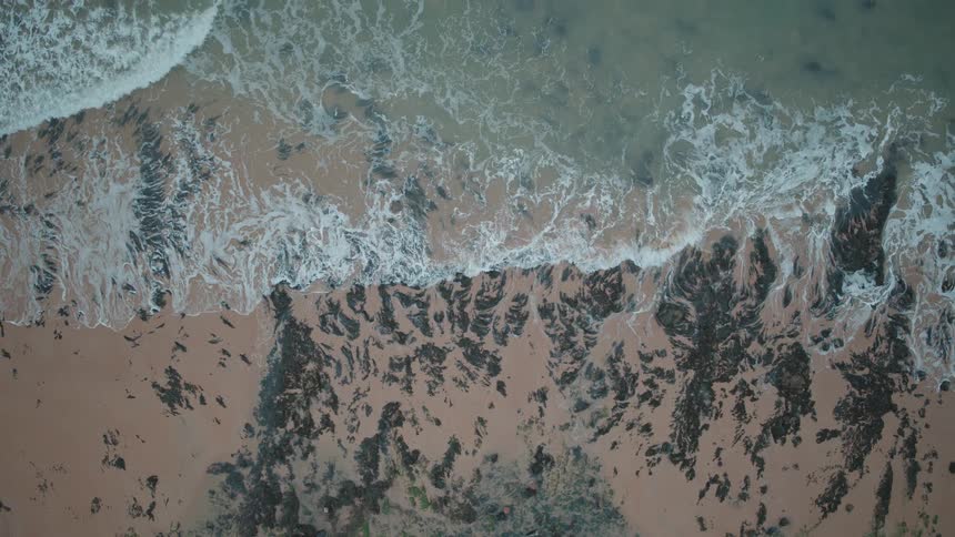 Mesmerizing aerial top-down slow-motion view of waves gently breaking on a rocky, sandy shoreline as the camera pulls up