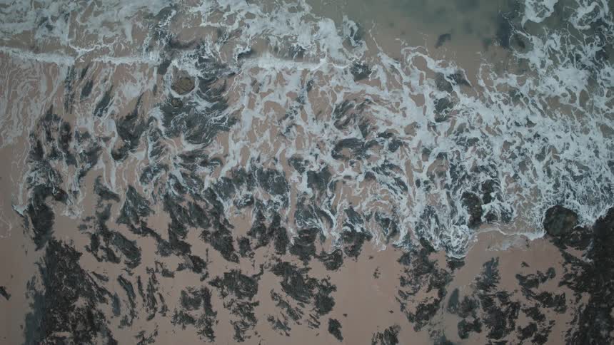 Slow-motion aerial shot from above captures the mesmerizing pattern of waves washing over a rocky coastline