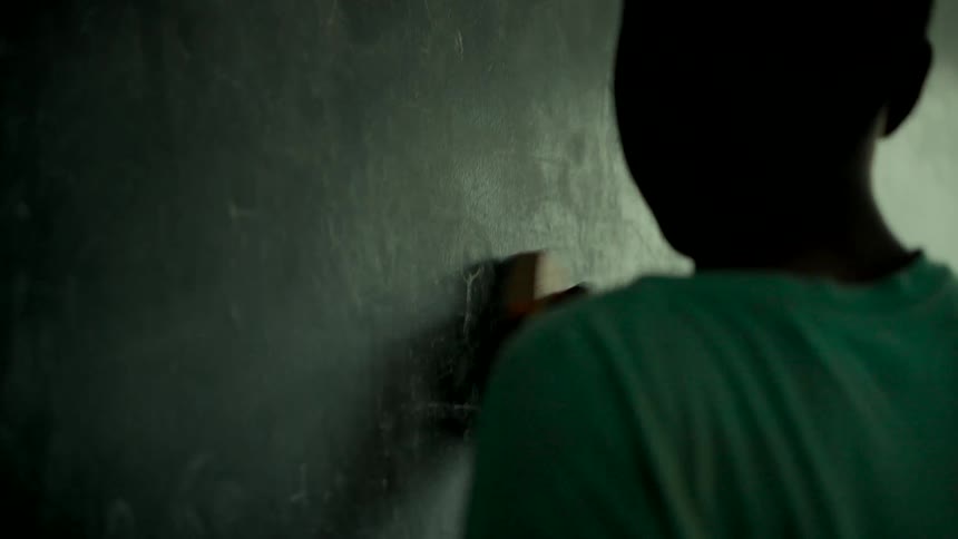 A student, seen from behind in silhouette, cleans a dusty chalkboard with an eraser in a dimly lit room