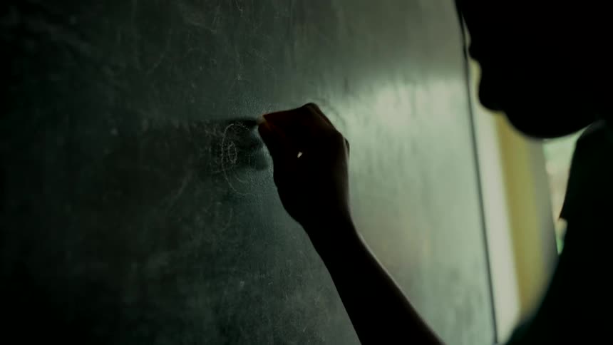 A silhouetted student's hand slowly draws with chalk on a dark chalkboard in a dimly lit classroom