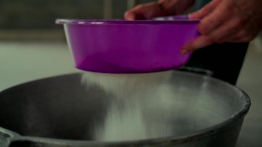Close-up of hands sifting flour from a purple plastic sieve into a large metal pot