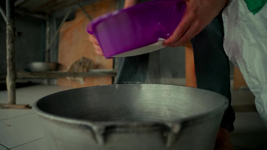 A person sifts white sugar through a purple sieve into a large metal cooking pot in slow motion