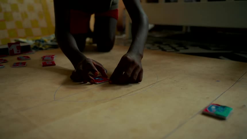 A close-up of a child's hands carefully moving a homemade game piece along a hand-drawn path on a wooden floor