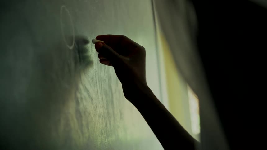 In a dimly lit classroom, a student's silhouetted hand practices writing on a blackboard with chalk