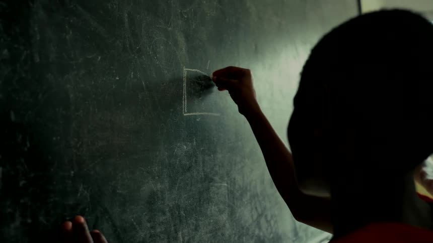 A silhouetted child carefully draws a square with white chalk on a dark chalkboard in a classroom