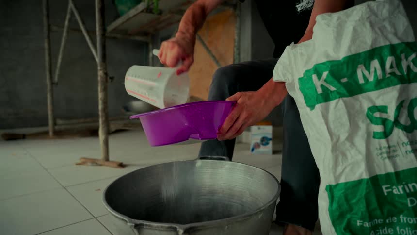 A man carefully measures and sifts flour into a large metal cooking pot in slow motion