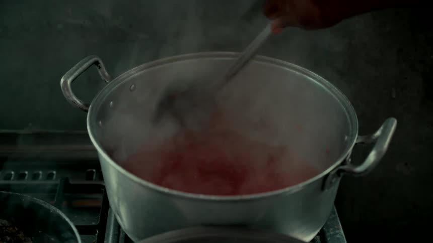 A cook stirs a simmering red stew in a large aluminum pot on the stove, releasing clouds of hot steam